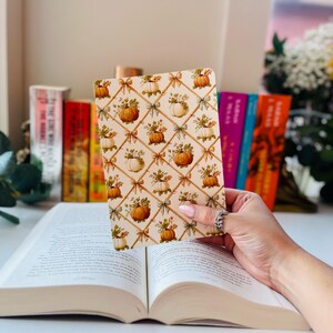 May include: A decorative notebook with a fall-themed pattern of pumpkins and floral arrangements. The notebook is held over an open book, with a background of colorful books and flowers. The notebook's design features a grid of pumpkins and bows.