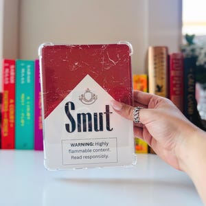 May include: Clear protective case with a red and white design resembling a cigarette pack. The word "Smut" is printed in bold black letters, with a warning label below. The case is held in front of a blurred background of books.