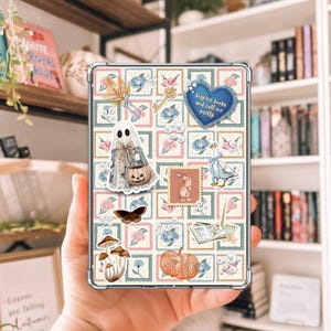May include: A clear phone case featuring a collage of whimsical illustrations. The design includes a ghost with a pumpkin, a key, a butterfly, mushrooms, and a pumpkin. The case has a blue heart with the text "buy me books and call me pretty".
