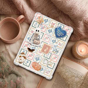 May include: A clear tablet case decorated with stickers, including a ghost, butterfly, and pumpkin. The case has a floral design with the text "buy me books and call me pretty" on a blue heart. A lit candle and a mug are also visible.