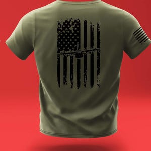 May include: A green t-shirt with a black and white distressed American flag design featuring a silhouette of a plane. The flag is centered on the back of the shirt.