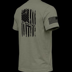 May include: An olive green short-sleeve t-shirt featuring a black distressed American flag graphic on the back with a silhouette of a cargo aircraft centered over the stripes. A small black American flag is printed on the right sleeve.