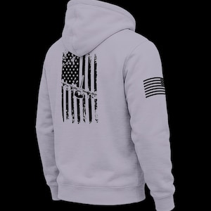 May include: A light gray hooded sweatshirt featuring a black distressed graphic on the back depicting an American flag with a military transport aircraft silhouette overlaid. A small black American flag graphic is printed on the right sleeve.