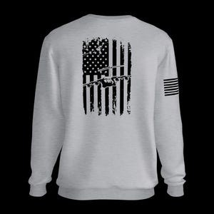Puede incluir: Una sudadera gris claro de cuello redondo con un gráfico negro desgastado de la bandera estadounidense en la espalda y la silueta de un avión de carga militar sobre las franjas. Un pequeño gráfico de bandera negra aparece en la manga derecha.