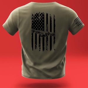 May include: A green t-shirt with a distressed black American flag design featuring a silhouette of a military aircraft. The flag is centered on the back of the shirt.