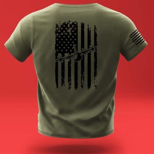 Puede incluir: Una camiseta verde con un diseño de bandera estadounidense desgastada en negro que presenta la silueta de una aeronave militar. La bandera está centrada en la parte posterior de la camiseta.