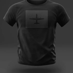 May include: Black t-shirt featuring a square design with an airplane silhouette. The design is a darker shade of black than the shirt itself. The shirt has short sleeves and a crew neck.