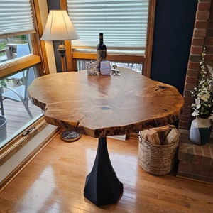 May include: A large, round, wooden table with a live edge top and a black pedestal base. The table is in a room with a window and a fireplace.