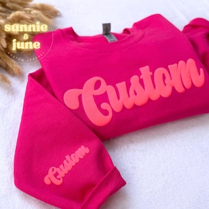 Puede incluir: Una sudadera rosa con la palabra "Custom" en una fuente retro grande de color rosa brillante en la parte delantera. La palabra "Custom" tambi&eacute;n est&aacute; en la manga de la sudadera.