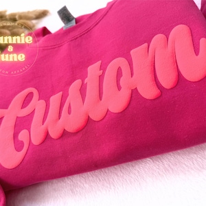 Puede incluir: Una sudadera rosa con la palabra "Custom" en una fuente grande, rosa brillante, de estilo retro.
