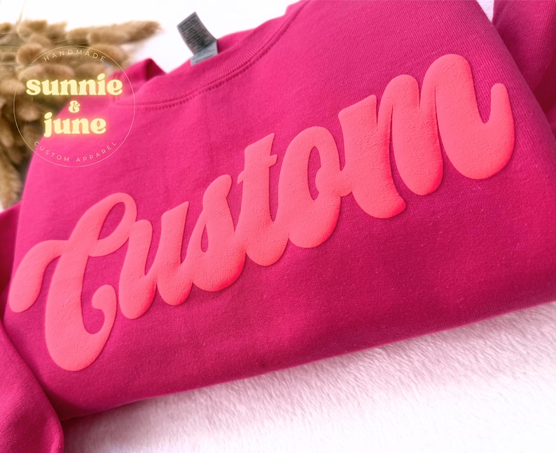 Puede incluir: Una sudadera rosa con la palabra "Custom" en una fuente grande, rosa brillante, de estilo retro.