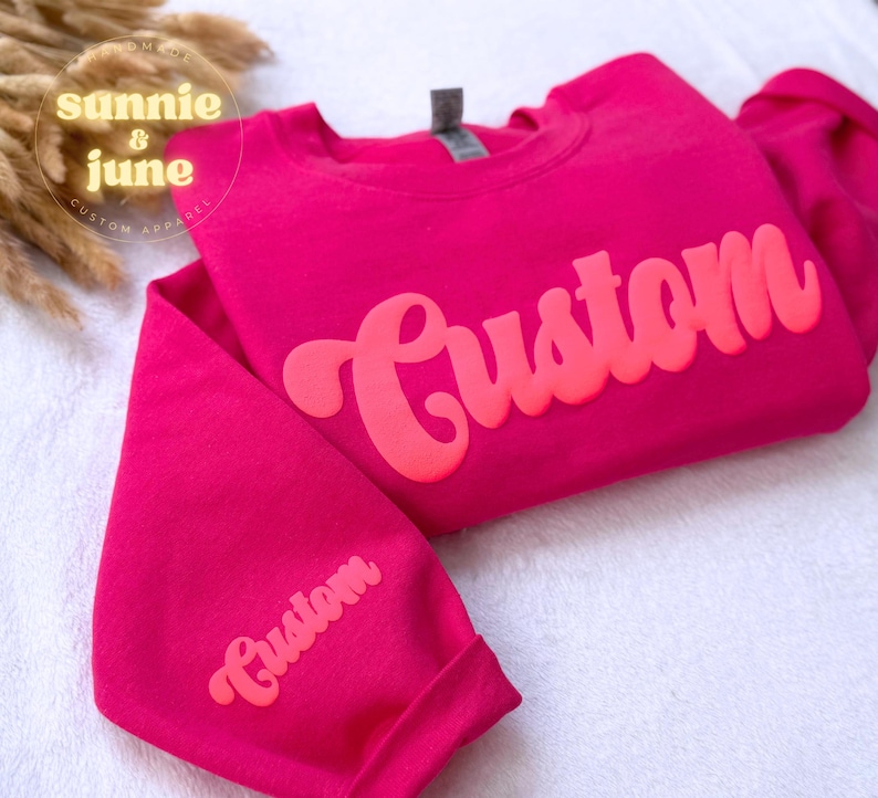 Puede incluir: Una sudadera rosa con la palabra "Custom" en una fuente retro grande de color rosa brillante en la parte delantera. La palabra "Custom" tambi&eacute;n est&aacute; en la manga de la sudadera.