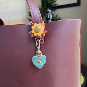 May include: A burgundy leather tote bag with brown handles. A decorative flower and heart charm hangs from the handle. The flower is yellow and brown, and the heart is teal. The bag is a simple, classic design.