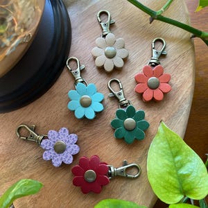 May include: Six colorful leather flower keychains with gold-tone metal clasps. The keychains are arranged on a wooden surface with green leaves.