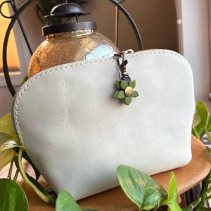 May include: Off-white leather cosmetic bag with a zipper closure. The bag features a decorative green flower charm with a gold center. The bag is sitting on a wooden surface, surrounded by green plants.