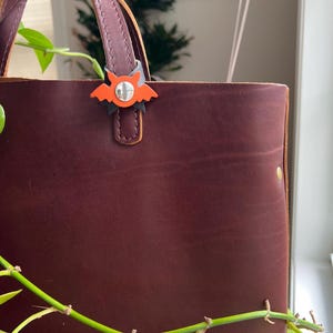 May include: A burgundy leather tote bag with a brown handle and a decorative orange bat charm. The bag has a simple, rectangular design with a sturdy handle. The bat charm is attached to the handle with a silver rivet.