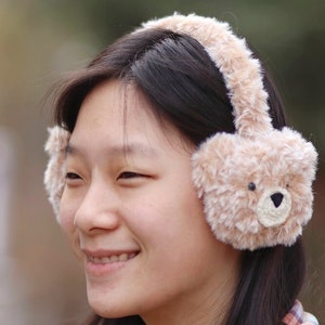 May include: A young woman with long dark hair is wearing a pair of brown fuzzy earmuffs shaped like a teddy bear. The earmuffs have a brown fur texture and a black nose.