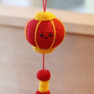 Può includere: Una lanterna di uncinetto rossa e gialla con un viso sorridente e un fiocco rosso.