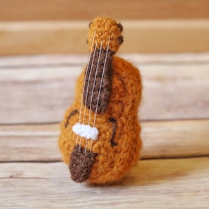 Puede incluir: Un pequeño violín de crochet hecho de hilo marrón y naranja con cuerdas blancas. El violín está sobre una superficie de madera.