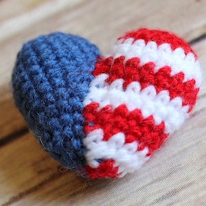 May include: A crocheted heart-shaped ornament in red, white, and blue. The heart is made of yarn and has a textured surface.