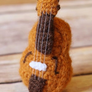 Puede incluir: Un violonchelo de juguete de crochet marr&oacute;n y blanco con cuerdas blancas. El violonchelo est&aacute; sobre una superficie de madera.