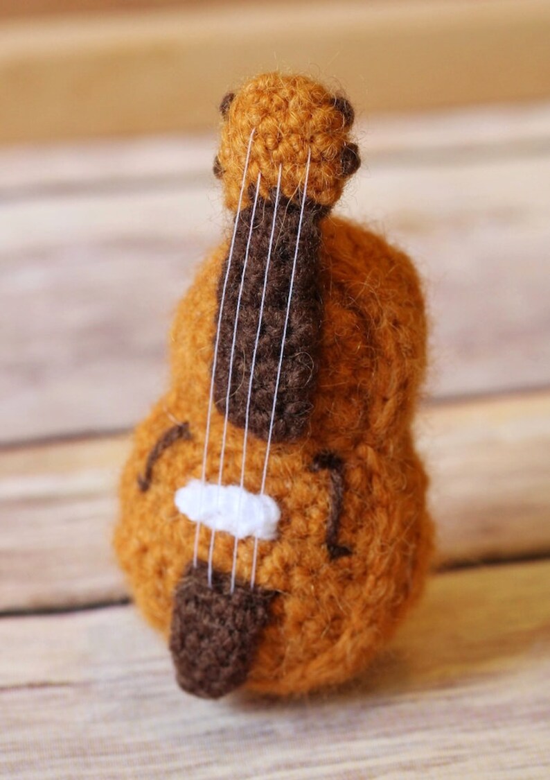 Puede incluir: Un violonchelo de juguete de crochet marr&oacute;n y blanco con cuerdas blancas. El violonchelo est&aacute; sobre una superficie de madera.