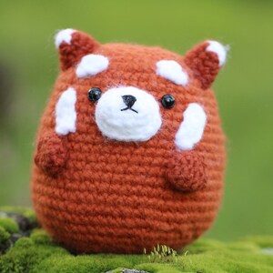 Pode incluir: Um brinquedo de pelúcia de panda vermelho de crochê com marcas brancas ao redor dos olhos e um nariz preto. O brinquedo está sentado em um leito de musgo verde.