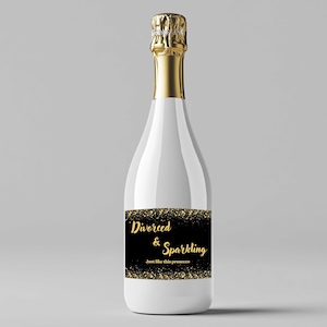 Könnte beinhalten: Eine weiße Champagnerflasche mit goldenem Folienverschluss und einem schwarzen Etikett. Auf dem Etikett steht "Divorced & Sparkling" in goldener Schrift, darunter der Satz "Just like this prosecco". Das Etikett hat einen goldenen Glitzerrahmen.