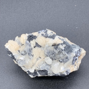 May include: A cluster of white and clear crystals on a dark gray rock. The crystals are arranged in a random pattern and have a shiny, translucent appearance.