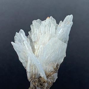 Peut inclure: Un groupe de cristaux de barytine blancs translucides en forme d'éventail. Les cristaux sont disposés en un motif radial, créant un affichage délicat et complexe. Les cristaux de barytine sont placés sur un fond sombre.