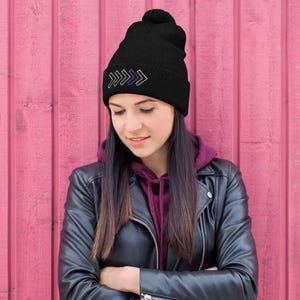 May include: Black beanie with a pom-pom on top. The beanie features a design of three stacked arrows in white, purple, and white. The hat is made of a soft, knitted material. The beanie is a stylish accessory for cold weather.