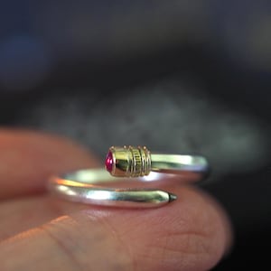May include: A silver bypass ring shaped like a pencil, featuring a metallic gold-toned ferrule and a small, vibrant pink gemstone at the tip, with a dark grey point at the opposite end of the band, creating a distinct writing instrument aesthetic.