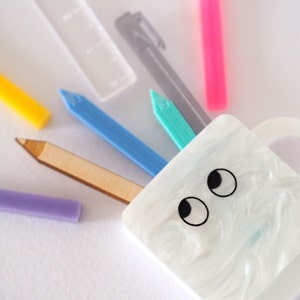 May include: A white ceramic mug with two black eyes painted on the front. The mug is filled with various colorful stationery items, including a blue pencil, a turquoise pencil, a wooden pencil, a yellow crayon, a purple crayon, a gray pen, and a pink highlighter.