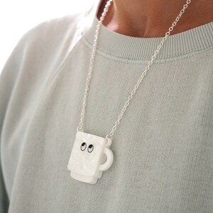 May include: A white ceramic mug necklace with black eyes. The mug is hanging from a silver chain.