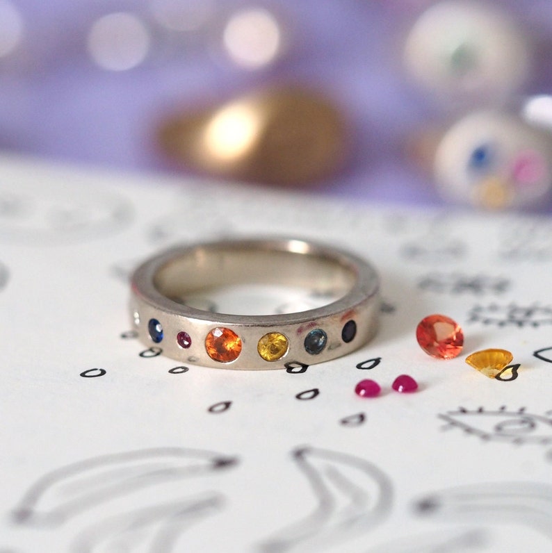 May include: A silver ring with a series of colorful gemstones, including blue, red, orange, and yellow. The ring is set against a white surface with scattered gemstones and a blurred background.
