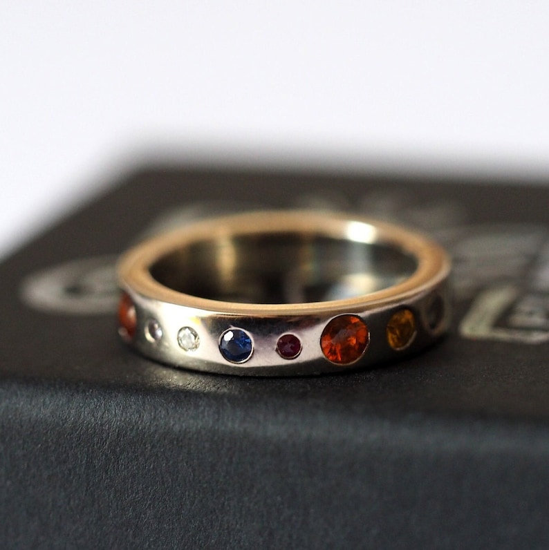 May include: A silver ring with a series of colorful gemstones. The ring features a variety of round-cut stones in shades of red, orange, yellow, blue, and white. The gemstones are set in a band of polished silver.