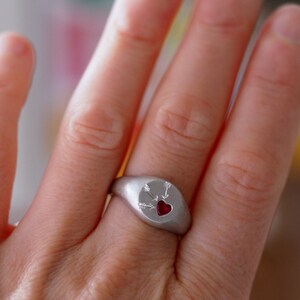 Puede incluir: Un anillo de sello de plata con un coraz&oacute;n rojo atravesado por tres flechas. El anillo se lleva en un dedo.