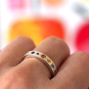 May include: A silver ring with a row of colorful gemstones. The gemstones are in various colors, including blue, red, orange, and yellow. The ring is on a finger, with a blurred, colorful background.