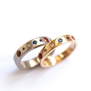 May include: Two rings, one silver and one gold, are displayed against a white background. Both rings are adorned with a series of colorful gemstones, including blue, red, and clear stones. The gemstones are evenly spaced around the bands.