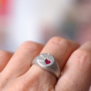Puede incluir: Un anillo de sello de plata con una piedra preciosa de rub&iacute; en forma de coraz&oacute;n engastada en el centro. El anillo est&aacute; grabado con un dise&ntilde;o de flechas que apuntan hacia el coraz&oacute;n.