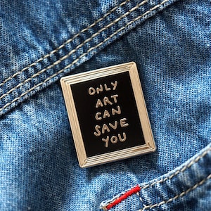 以下が含まれることがあります： 黒い背景に金色の文字で「Only Art Can Save You」と書かれた、銀色の枠のついたエナメルピンです。
