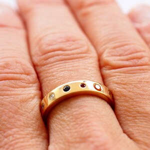 May include: A gold band ring with alternating gemstones. The ring features a series of small, round gemstones in various colors, including black, red, and clear. The ring is worn on a finger, showcasing its design and craftsmanship.