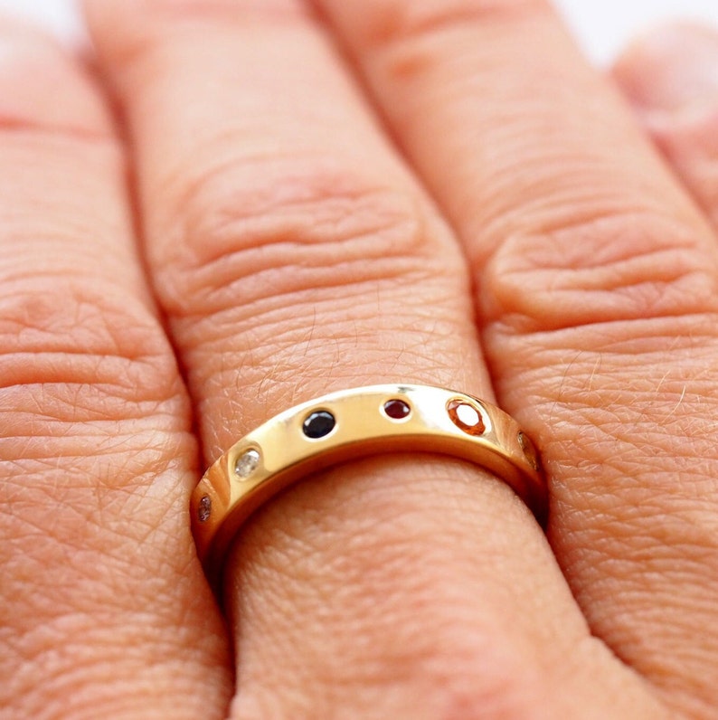 May include: A gold band ring with alternating gemstones. The ring features a series of small, round gemstones in various colors, including black, red, and clear. The ring is worn on a finger, showcasing its design and craftsmanship.