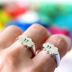 Może przedstawiać: Dwa srebrne pierścionki, każdy z chmurkowym wzorem i uśmiechniętą twarzą. Pierścionki są noszone na palcach, na rozmytym, kolorowym tle. Wzór chmurki jest w jasnym srebrnym kolorze.