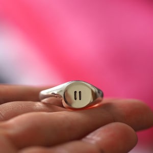 May include: A silver signet ring with a rounded face featuring two vertical lines. The ring is held in a hand against a pink background. The ring's design is simple and modern.
