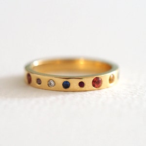 May include: A gold ring with a series of colorful gemstones. The ring features a diamond and various other gemstones in red, blue, and yellow. The ring is set against a white background.