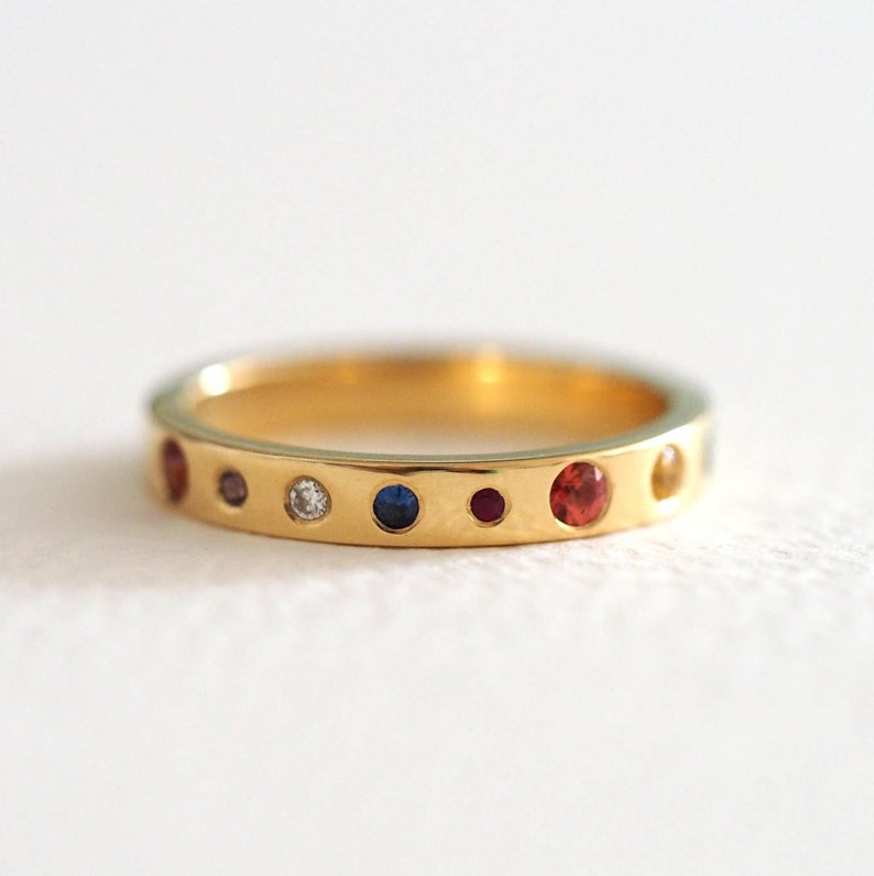 May include: A gold ring with a series of colorful gemstones. The ring features a diamond and various other gemstones in red, blue, and yellow. The ring is set against a white background.