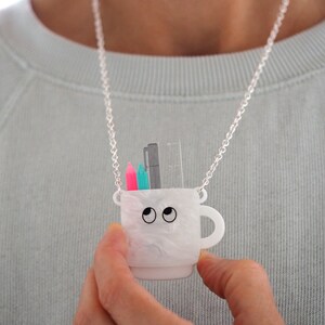 May include: A white ceramic mug necklace with a silver chain. The mug has googly eyes and holds a ruler, a black pen, and two colored pencils.