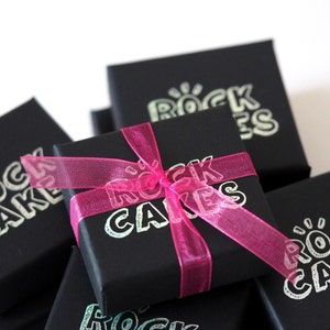 May include: Black gift boxes with the text "ROCK CAKES" in a silver font. A bright pink ribbon is tied around one box in a bow. The boxes are stacked on top of each other.