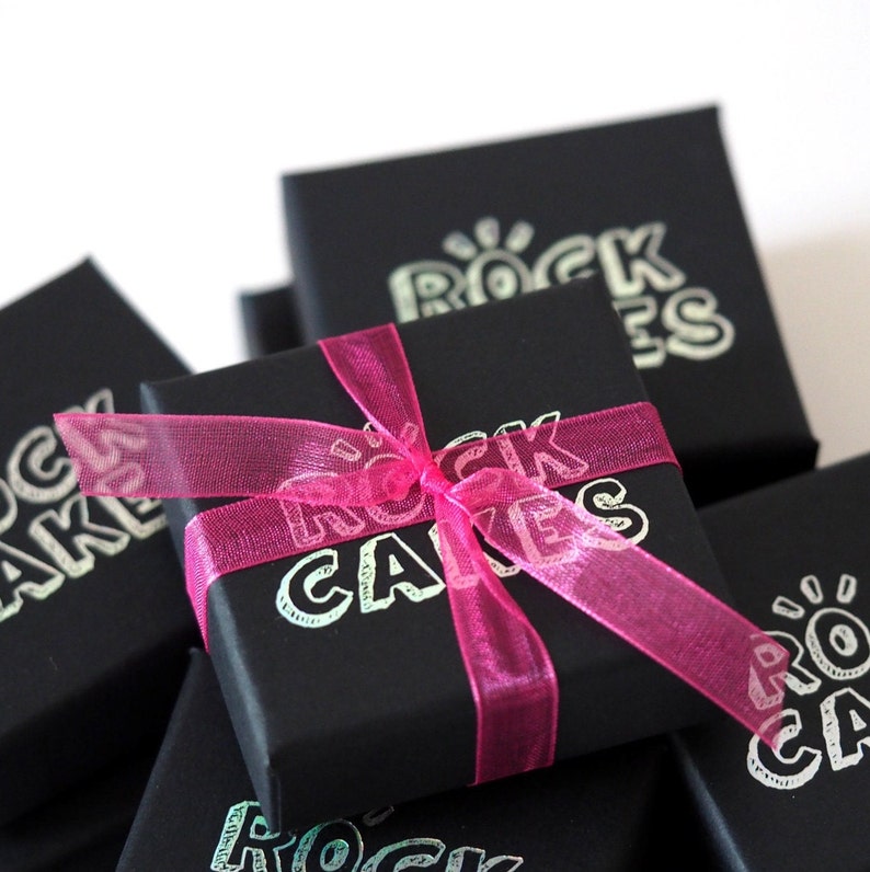 May include: Black gift boxes with the text "ROCK CAKES" in a silver font. A bright pink ribbon is tied around one box in a bow. The boxes are stacked on top of each other.